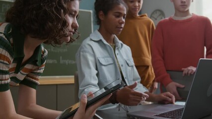 Medium shot of 14-year-old African American schoolgirl showing and explaining coding project on laptop screen to group of multiethnic classmates, standing around desk at programming lesson - Powered by Adobe