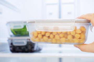 A female hand holds a glass box with chickpeas against the background of an open refrigerator, close-up.