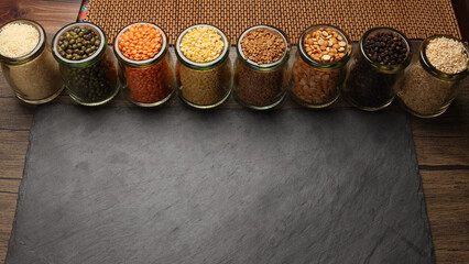 Variety of grains red green yellow dal rice sesame methi legumes in glass jars displayed on a dark black slate surface copy text space