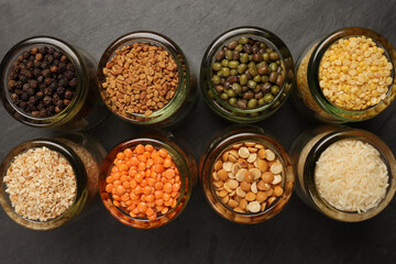 Variety of grains red green yellow dal rice sesame methi legumes in glass jars displayed on a dark black slate surface copy text space