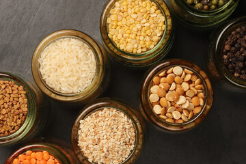 Variety of grains red green yellow dal rice sesame methi legumes in glass jars displayed on a dark black slate surface copy text space