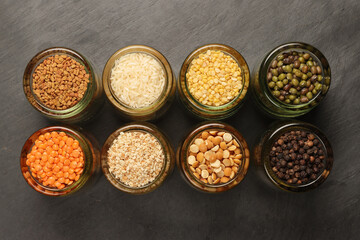Variety of grains red green yellow dal rice sesame methi legumes in glass jars displayed on a dark black slate surface copy text space