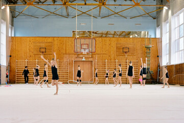 Obraz premium Group of Caucasian girls practicing rhythmic gymnastics with hoops in sports hall, performing synchronized movements on gymnasium floor, some raising arms, others spinning hoops