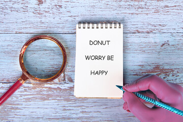 A hand writes a note saying DONUT WORRY BE HAPPY near a magnifying glass. It sits on a light wooden desk suggesting a positive message