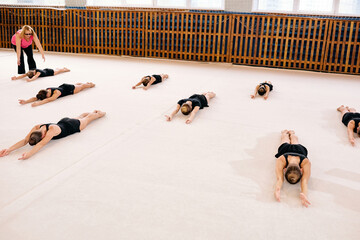 Group of Caucasian girls lying on gym floor stretching arms forward during gymnastics class, female coach supervising from side, all participants wearing athletic outfits, indoor sports facility