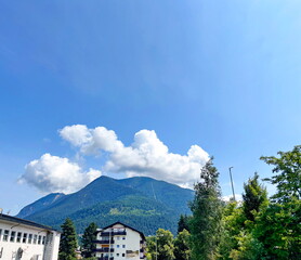 View from the street of a mountain town on the surrounding mountains whose tops touch the snow-white clouds in the blue sky.