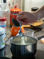 Preparing milk porridge. A person's hand pours semolina, oatmeal or rice cereal into a pan with milk with a spoon. Preparing breakfast.