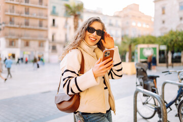 Fototapeta premium A young woman in sunglasses, holding phone, strolls through old streets of the city on sunny day. A beautiful tourist enjoys sunset, blogs, and texts on the phone. Concept of tourism, weekends.