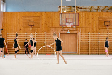 Obraz premium Caucasian girls practicing rhythmic gymnastics with hoops in gymnasium, young athletes performing exercises on mat, one girl standing in foreground holding hoop, others training behind