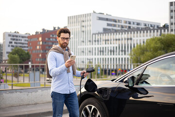 Naklejka premium A man checks his phone while his electric vehicle charges in an urban setting.
