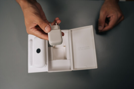Top view of young man unpacking holding white smartphone charger being unpacked from cardboard box, sitting on gray table. Concept of contemporary gadget unboxing. - Powered by Adobe