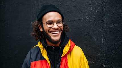 Man wearing glasses, smiling, standing against a black wall, wearing a yellow jacket with red sleeves, and a black beanie.