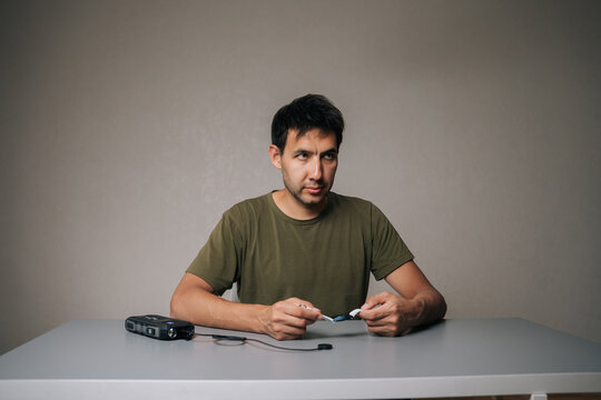 Studio portrait of young man sitting at gray table, preparing charging smartwatch with portable charger, looking away, surrounded by minimalist gray background, blending technology, modern lifestyle.