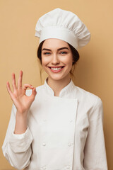 Joyful female chef posing with an OK sign in her kitchen uniform against a beige backdrop