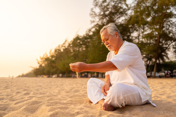Asian senior man hand holding sand during practicing meditation on the beach at summer sunset. Retirement elderly people do outdoor relaxing exercise. Mental health care and motivation concept.