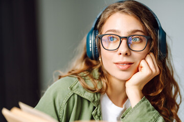 Close-up of young woman wearing wireless headphones and reading book in cozy room. Beautiful woman...