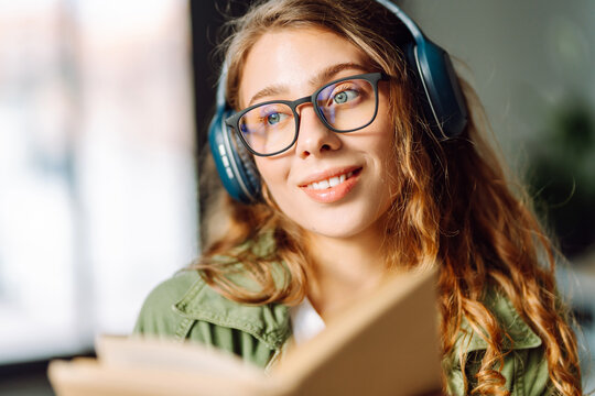 Close-up of young woman wearing wireless headphones and reading book in cozy room. Beautiful woman enjoys reading book and listening to music in cozy atmosphere. Concept of relaxation and enjoyment.