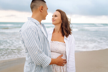 A young couple expecting a child stands on the beach and enjoys the seascape at sunrise. The pregnant woman and her husband spend time together. Concepts of family, love, and nature.