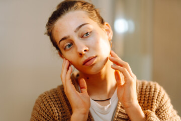 Portrait of young woman examining  face in mirror while standing in bathroom. Woman touches skin...