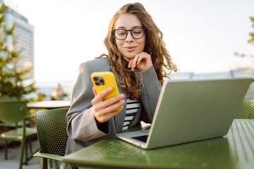 Woman with phone sits on terrace of street cafe with laptop at sunset. Beautiful female freelancer works on laptop and uses phone while enjoying sunny day on rooftop. Concept of technology, blogging.