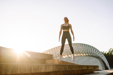 An athletic woman works out on a special platform at sunset. A beautiful woman in athletic wear performs jumping jacks, warming up before running outdoors. Active lifestyle.