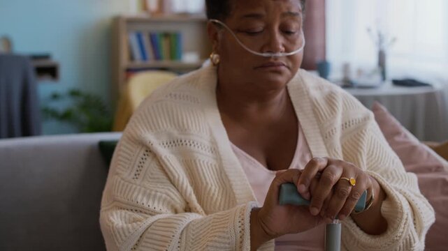 Tilt up portrait of sick senior lady with nasal cannula and crutch looking at camera while sitting on couch in living room