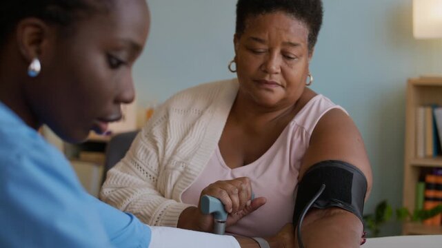 Tilt up shot of medical specialist in uniform measuring blood pressure of mature woman with tonometer indoors