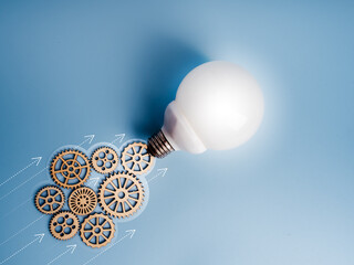 White globe light bulb with gear wheels as jet smoke and forward arrows on blue background. Innovation, clean energy, sustainable power, business growth, future-driven technological progress concept.