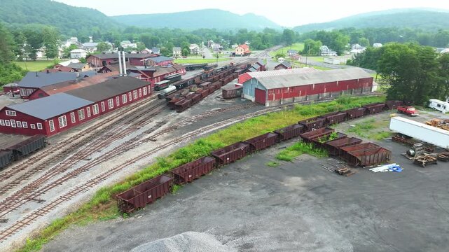 This clip showcases an aerial perspective of a train yard in a small town. Old train tracks run through the area, with storage containers, wood piles, and buildings visible in the background.