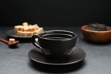 Aromatic tea in cup, brown sugar and dried leaves on black table, closeup