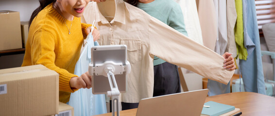 Two young asian woman live online clothing store while showing clothing with tablet on social at...