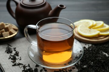 Aromatic black tea in glass cup, dried leaves, brown sugar, lemon and teapot on table, closeup