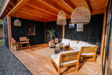 Stylish outdoor lounge with wooden furniture, white cushions, wicker pendant lights, black wood paneling, and cozy decor in a modern cabin setting.