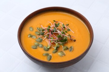 Delicious pumpkin soup with seeds and microgreens in bowl on white tiled table, closeup