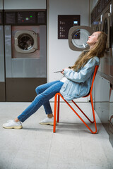 Woman listening to music with smartphone while waiting laundry in self service laundromat