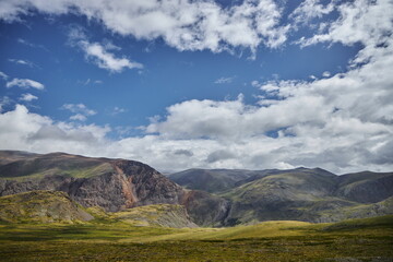 Vast green hills stretch across the foreground while majestic mountains rise in the background. Sky is bright with fluffy clouds, creating a tranquil midday scene in nature. Russia, Altai Mountains
