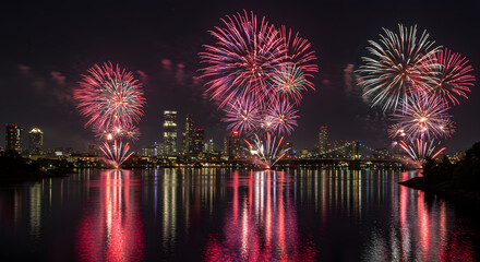 Fototapeta premium Fireworks bursting over city skyline with reflections on river below