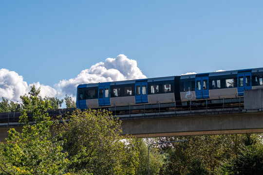 Blue train crossing elevated railway over trees under clear sky, showing integration of urban transport and surrounding natural landscape in Stockholm
