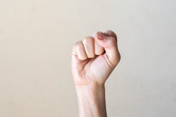 A raised fist with a light skin tone against a plain beige background. The hand is clenched,...