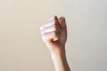 A raised fist with a light skin tone against a plain beige background. The hand is clenched,...