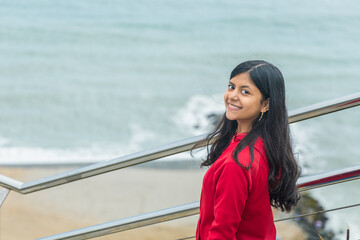 A teen in a red jacket stands on a beach, smiling