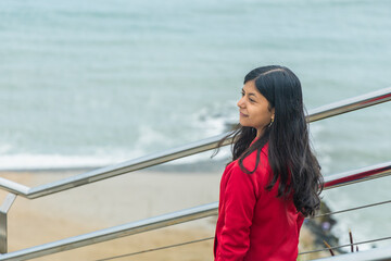 A teen in a red jacket stands on a metal railing overlooking the ocean