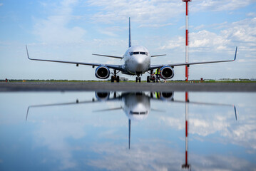 A large commercial airplane is parked on the tarmac at an airport, with its reflection visible on the wet surface. The aircraft is positioned in front of a control tower, and the sky is partly cloudy.