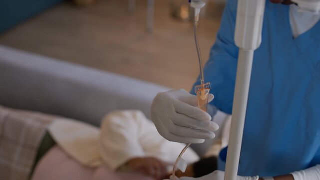 Tilt down shot of African American female medical worker in scrubs adjusting IV drip tube for patient resting on sofa at home