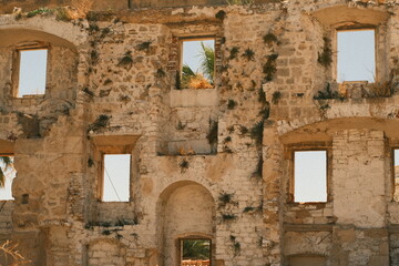 Facade of an old ruined stone building with empty window frames and plants growing on the walls. Historic Mediterranean architecture and cultural heritage.