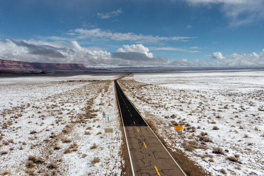 Aerial view of a long, dark road cutting through a snow-dusted landscape, contrasting with the distant red rock formations under a bright, partly cloudy sky, Sedona, Arizona, United States.