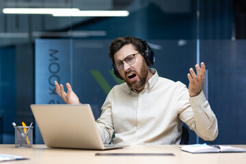 A young male office worker sits at his desk wearing headphones in front of a laptop and singing