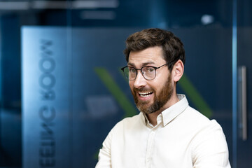 Close-up portrait of a smiling young man in glasses and shirt standing in the office, looking and smiling at the camera