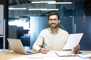 Portrait of a young smiling businessman working in the office at a laptop and with documents, looking at the camera, signing papers