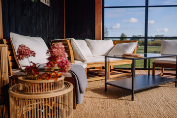 Close-up of a cozy outdoor seating area with white cushions, a glass-top table, pink flowers, and a scenic field view through large windows.